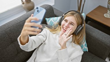 Kulaklık takan neşeli sarışın bir kadın oturma odasındaki kanepeden arıyor.