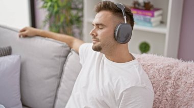 Rahatlamış İspanyol adam kulaklıkla müzik dinliyor evdeki bir kanepede oturuyor.