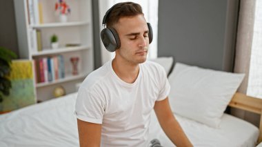 Huzurlu genç bir adam, rahat yatak odası ortamında kulaklıklarla müzik dinlemekten zevk alır. Rahatlık ve rahatlık yayar..