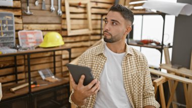 Sakallı genç bir adam, iyi donanımlı bir marangozluk atölyesinde bir tablet tutuyor..