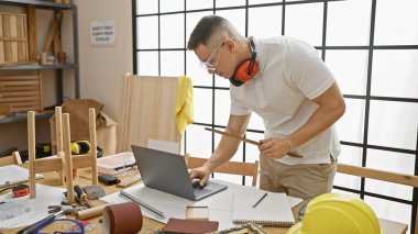 İyi organize edilmiş bir marangoz atölyesinde dizüstü bilgisayarla araştırma yapan İspanyol bir adam.