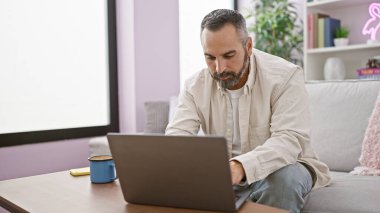 Olgun sakallı bir adam rahat bir oturma odasında dizüstü bilgisayarda çalışıyor. Kanepesi ve dekorasyonu var..