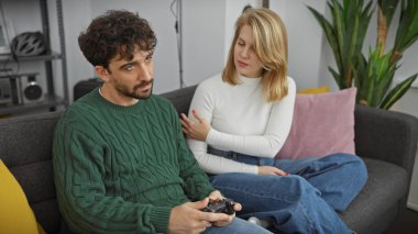 Sıcak bir oturma odasında, bir kadın video oyunu oynarken erkeğin omzuna dokunur, bu da ev hayatındaki bağlantıyı gösterir..