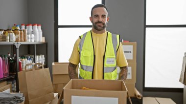 'Gönüllü' yeleği giyen sakallı genç bir İspanyol, kutularla dolu kapalı bir depoda duruyor..