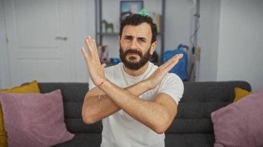Orta yaşlı, sakallı bir adam reddetme hareketi yapıyor ya da modern bir oturma odasında duruyor olumsuzluk ya da reddedilmeyi ifade ediyor..