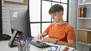 Genç beyaz bir erkek, modern bir üniversite kütüphanesinde bilgisayar başında özenle okuyor..