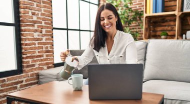 Genç ve güzel İspanyol kadın dizüstü bilgisayar kullanıyor. Evdeki fincana kahve döküyor.