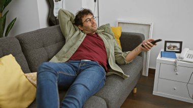 Gözlüklü ve sakallı sıradan bir İspanyol adam evdeki koltukta uzanıyor, elinde uzaktan kumandayla..