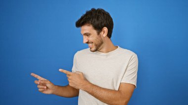 Yakışıklı, sakallı, mavi bir arka plana gülümseyen ve işaret eden genç bir İspanyol..