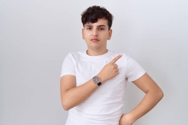 Gündelik beyaz tişört giyen, el parmaklarıyla kenarı gösteren, ciddi ve sakin bir yüz ifadesi olan genç, ikili olmayan bir adam. 