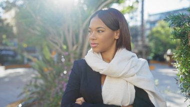 Confident black woman in business attire with a scarf crosses arms outdoors in a park setting. clipart