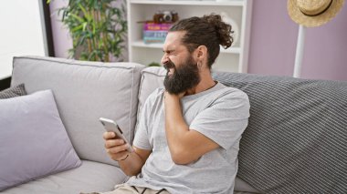 Evde kanepede akıllı telefon kullanırken boynu ağrıyan sakallı yakışıklı bir İspanyol..