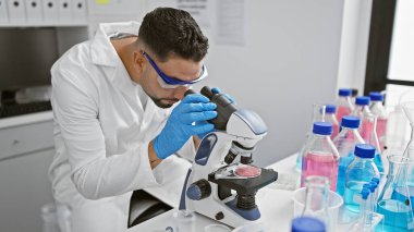 Genç bir adam, bilimsel ekipmanlarla çevrili bir laboratuvar ortamında mikroskop altında bir numuneyi inceliyor..