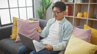 Modern bir apartman dairesinde renkli yastıkları olan Asyalı bir adam dizüstü bilgisayar kullanıyor.