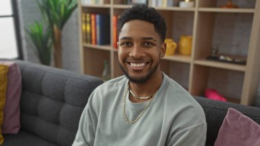 Gülümseyen Afrikalı Amerikalı adam kitap ve ev dekoruyla dolu modern bir oturma odasında rahatça oturuyordu.