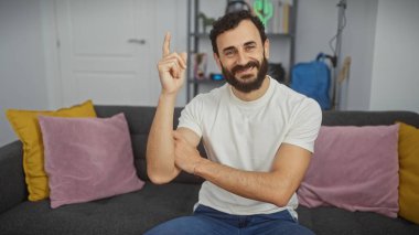 Mutlu sakallı bir adam, günlük kıyafetlerle kanepede otururken bir fikir belirtiyor..