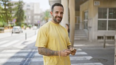 Sakallı, gülümseyen genç İspanyol bir adam elinde akıllı bir telefonla şehir yaşamını gözler önüne seriyor..