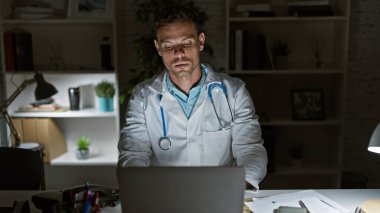 Laboratuvar önlüklü İspanyol adam geceleri klinikte dizüstü bilgisayarla çalışıyor.