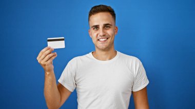 Yakışıklı İspanyol bir adam elinde kredi kartıyla mavi arka planda gülümsüyor..