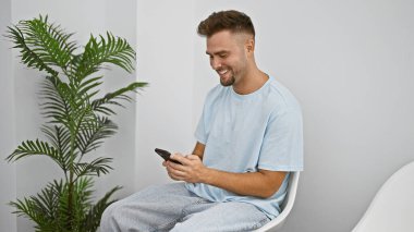 Modern beyaz bir odada yeşil bir bitkiyle otururken akıllı telefon kullanan gülümseyen genç adam.