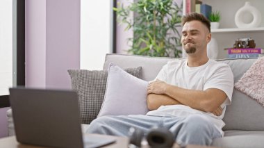 Modern bir oturma odasında dizüstü bilgisayarı ve kamerasıyla kanepede dinlenen İspanyol bir adam.