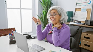 Gri saçlı neşeli bir kadın parlak bir ofis ortamında anahtar sunuyor, mesleki başarı ve memnuniyeti somutlaştırıyor..