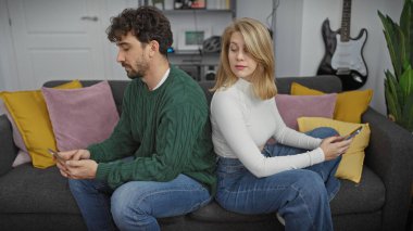 Bir kadın, renkli yastıklarla kanepede sırt sırta otururken bir erkeğin telefonuna gizlice bakar..