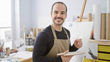 Resim stüdyosunda elinde fırça ve palet tutan sakallı, gülümseyen kel bir adam..