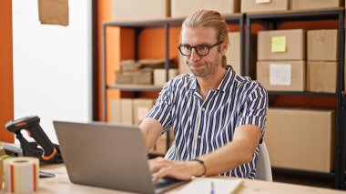 Gözlüklü bir adam, raflar ve kutularla çevrili modern bir depoda dizüstü bilgisayarda çalışıyor..