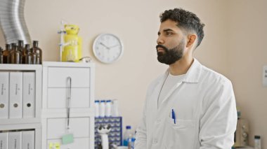 Raflar ve ekipmanlarla dolu bir tıp laboratuarında düşünceli, sakallı yakışıklı bir adam..