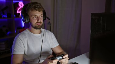 Gece karanlık bir odada sakallı ve kulaklıklı beyaz bir adam video oyunları oynuyor.