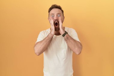 Sarı arka planda dikilen sakallı orta yaşlı adam korkmuş ve şok olmuş, şaşırmış ve şaşırmış bir yüz ifadesi var. 