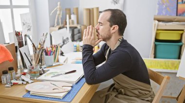 Önlük takan düşünceli kel bir adam resim malzemeleri ile çevrili bir sanat stüdyosunda oturuyor..