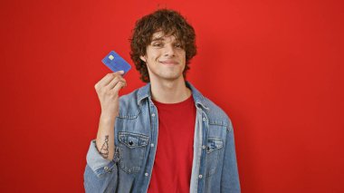 Kıvırcık saçlı, kırmızı arka planda kredi kartı tutan gülümseyen genç adam.