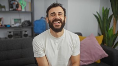 Sakallı adam neşeli bir şekilde modern bir oturma odasında gülüyor.