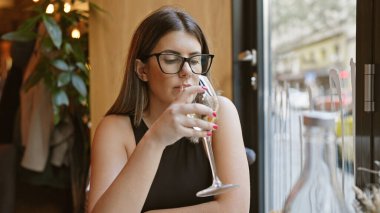 Esmer, gözlüklü bir kadın modern bir restoranda şarap yudumluyor, rahat bir yemek atmosferini somutlaştırıyor..