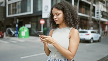 Çekici, genç İspanyol bir kadın. Ciddi bir konsantrasyon gösteriyor. Akıllı telefonuyla mesajlaşıyor, güneşli şehir caddesinde dikiliyor, havalı şehir yaşam tarzını yaşıyor..