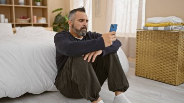 Olgun sakallı İspanyol adam yatak odasında oturuyor. Akıllı telefon kullanıyor, etrafı dekorasyonla çevrili..