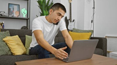 Latin kökenli genç bir adam, rahat bir oturma odasında, telefonda konuşurken dizüstü bilgisayarla birden fazla iş yapıyor..