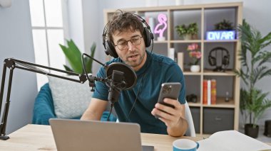 Stüdyoda sakallı İspanyol bir adam podcast yaparken akıllı telefon kullanıyor.