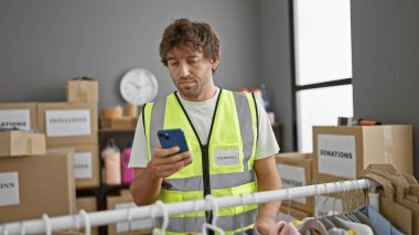 Gönüllü olarak etiketlenmiş yansıtıcı yelek giyen bir adam bağış merkezinin deposunda akıllı telefon kullanıyor..
