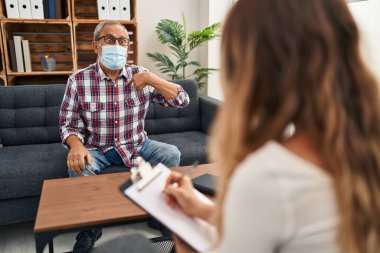 Beyaz saçlı neşeli bir son sınıf öğrencisi, bir psikoloji kliniğinde gülümserken ve maske takarken gururla kendini işaret ediyor, yoğun konsültasyonlar arasında iyimser davranışlar sergiliyor.
