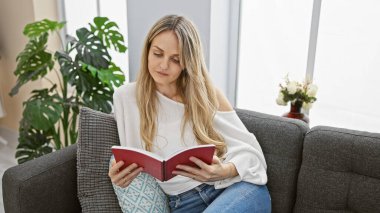 Huzurlu genç bir kadın, rahat rahat oturduğu gri bir kanepede kitap okuyor..