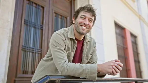 stock image Smiling, bearded hispanic man leaning on a railing with a blurred building facade in an urban setting.