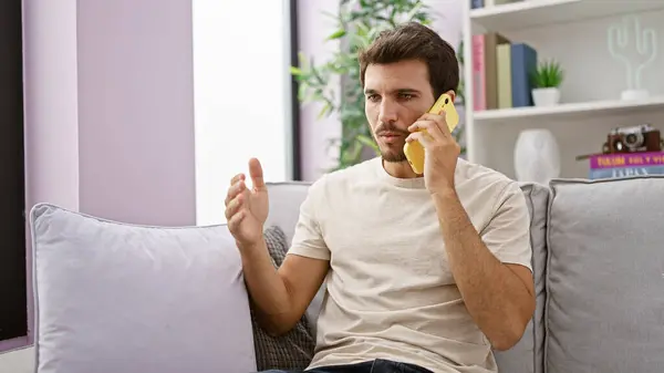 stock image A young hispanic man with a beard engages in conversation on a yellow phone while sitting on a grey couch indoors.