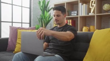 Sakallı genç bir İspanyol, modern bir apartmanın rahat oturma odasında dizüstü bilgisayarıyla çalışıyor..