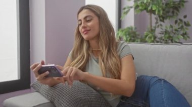 Sarışın genç bir kadın elinde akıllı bir telefon tutarken gülümsüyor, gri bir kanepede oturuyor..