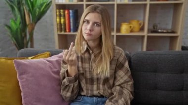 Angry young blonde woman sitting on sofa at home, showing the 'fuck you' middle finger sign, expressing a rude, impolite 'back off' concept