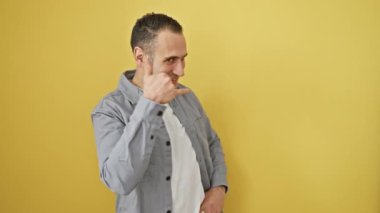 Gömlekli neşeli genç İspanyol adam sarı izole edilmiş arka plana karşı duruyor. Sırıtarak, parmaklarıyla ve eliyle bir telefon konuşması yapıyormuş gibi yapıyor. iletişim konsepti.