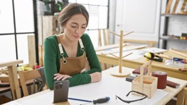 Bir kadın atölyede marangozluk aletleri, akıllı telefon ve koruyucu teçhizatla el işi yapıyor..